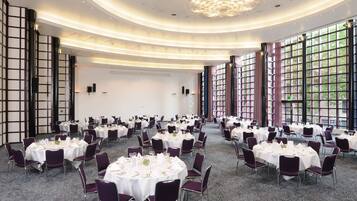 A large banquet hall with round tables set for dining, featuring a modern chandelier and large windows.