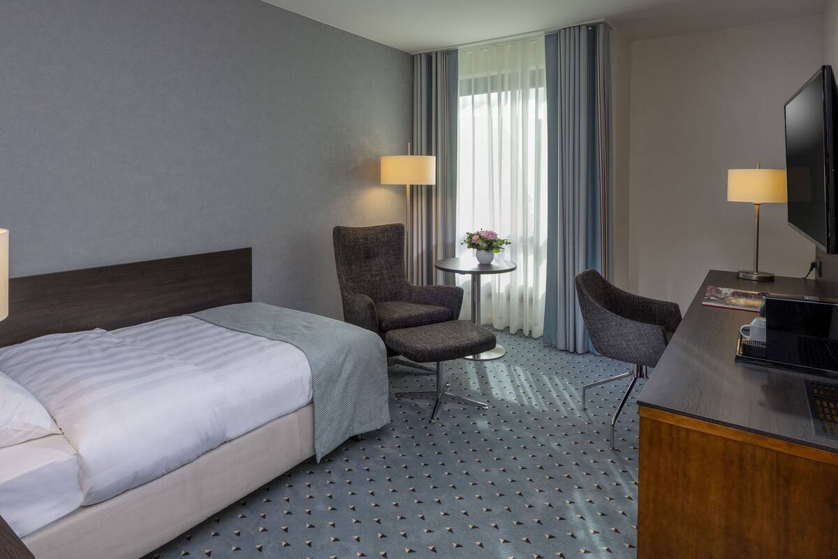 Ein Hotelzimmer mit Bett, zwei Sesseln, einem kleinen Tisch mit Lampe, einem Fernseher und einem Fenster mit Vorhängen.