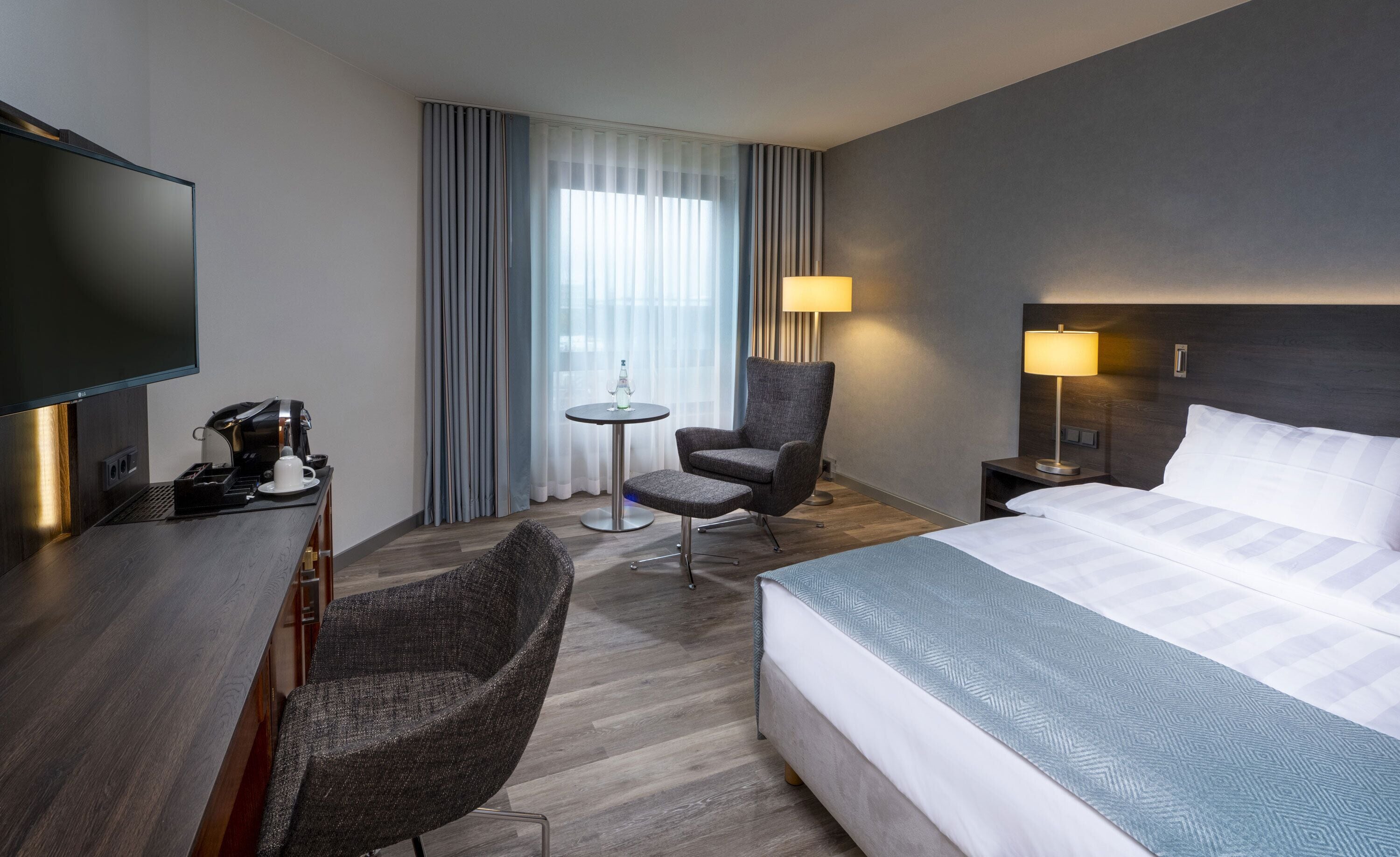 A hotel room with a bed, a desk with a coffee maker, a chair, a small table, a lamp, and a TV.