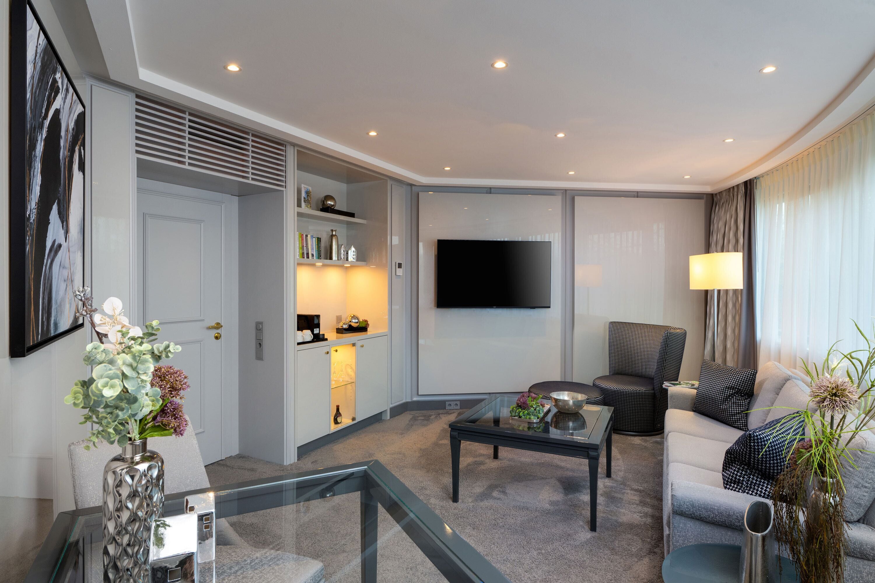 A modern living room with a glass coffee table, a sofa, a TV mounted on the wall, and a dining area with a kitchen counter and shelves.