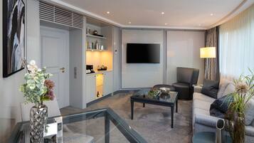 A modern living room with a glass coffee table, a sofa, a TV mounted on the wall, and a dining area with a kitchen counter and shelves.