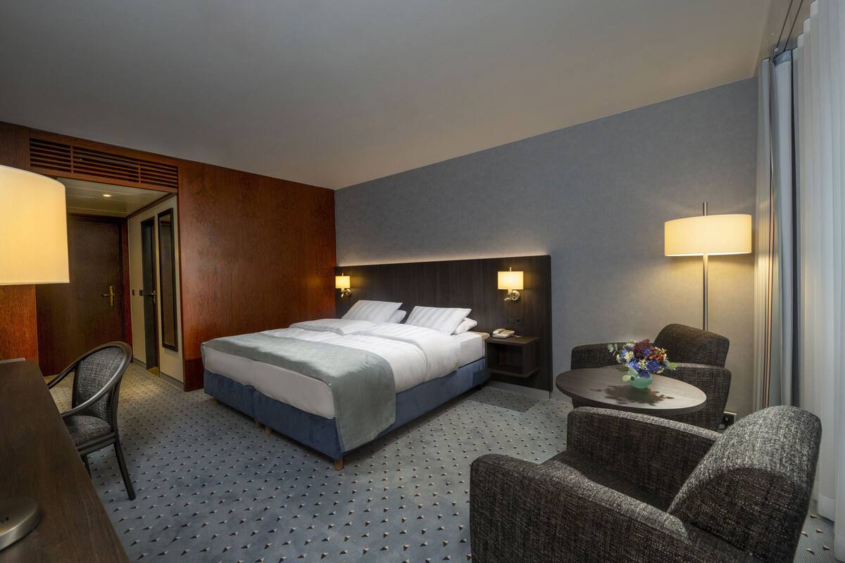 Ein Hotelzimmer mit einem Bett, zwei Nachttischen, einem Schreibtisch, einem Sessel und einem kleinen Tisch mit Blumen in einer Vase.