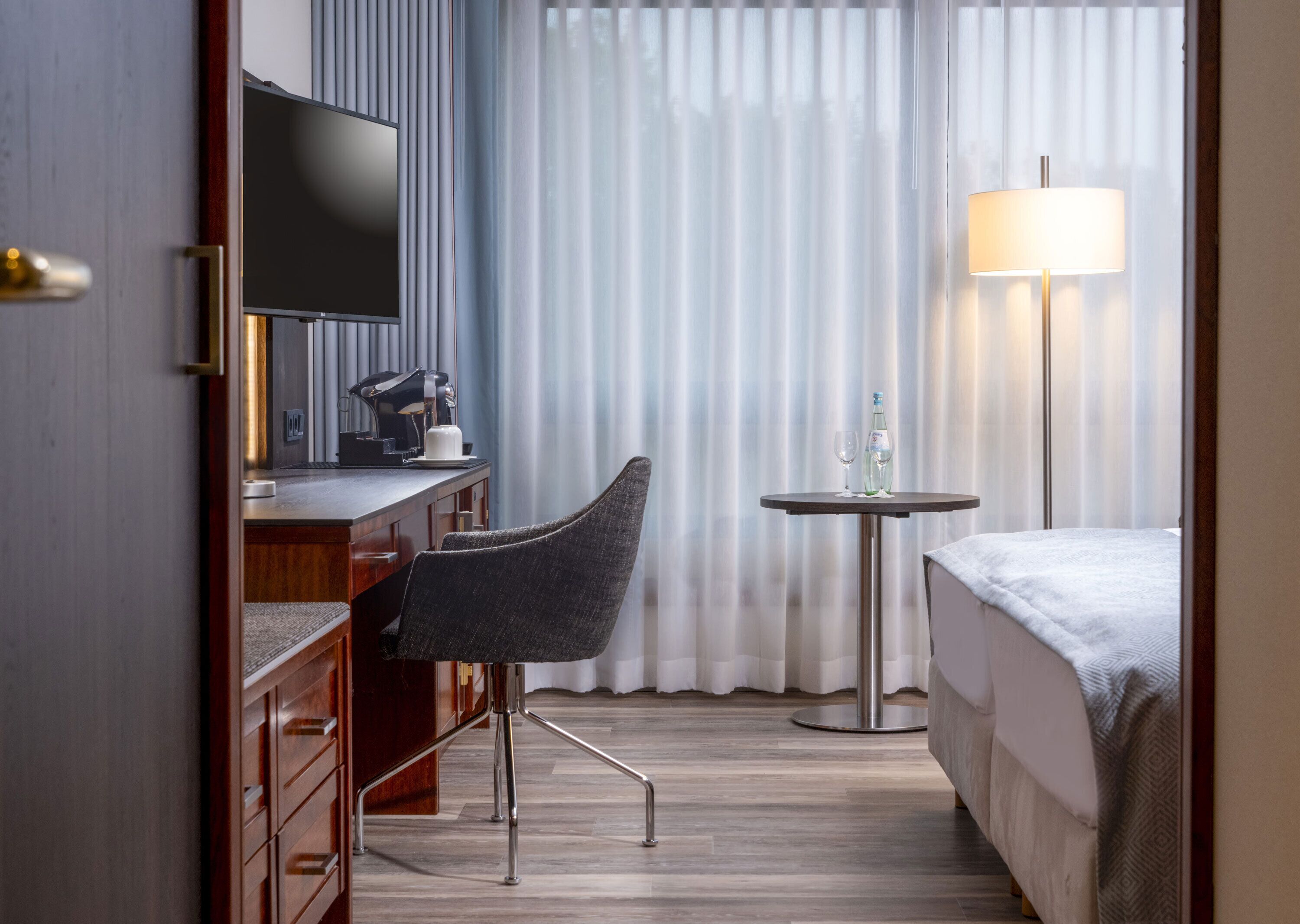 A hotel room with a bed, a desk with a chair, a television, a lamp, and a window with curtains.