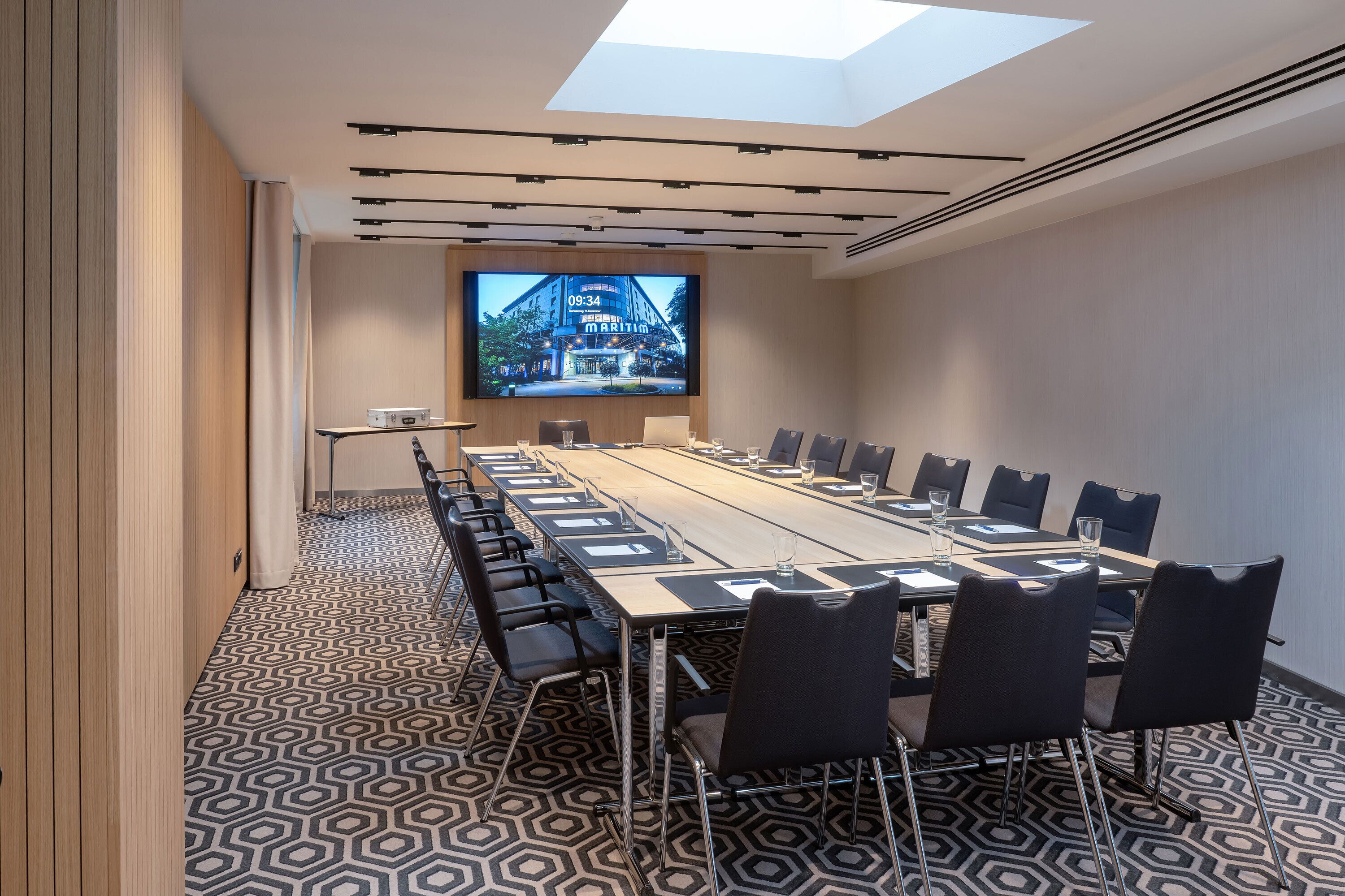 Ein Konferenzraum mit einem langen Tisch, Stühlen, einer großen Leinwand und einem Musterboden.