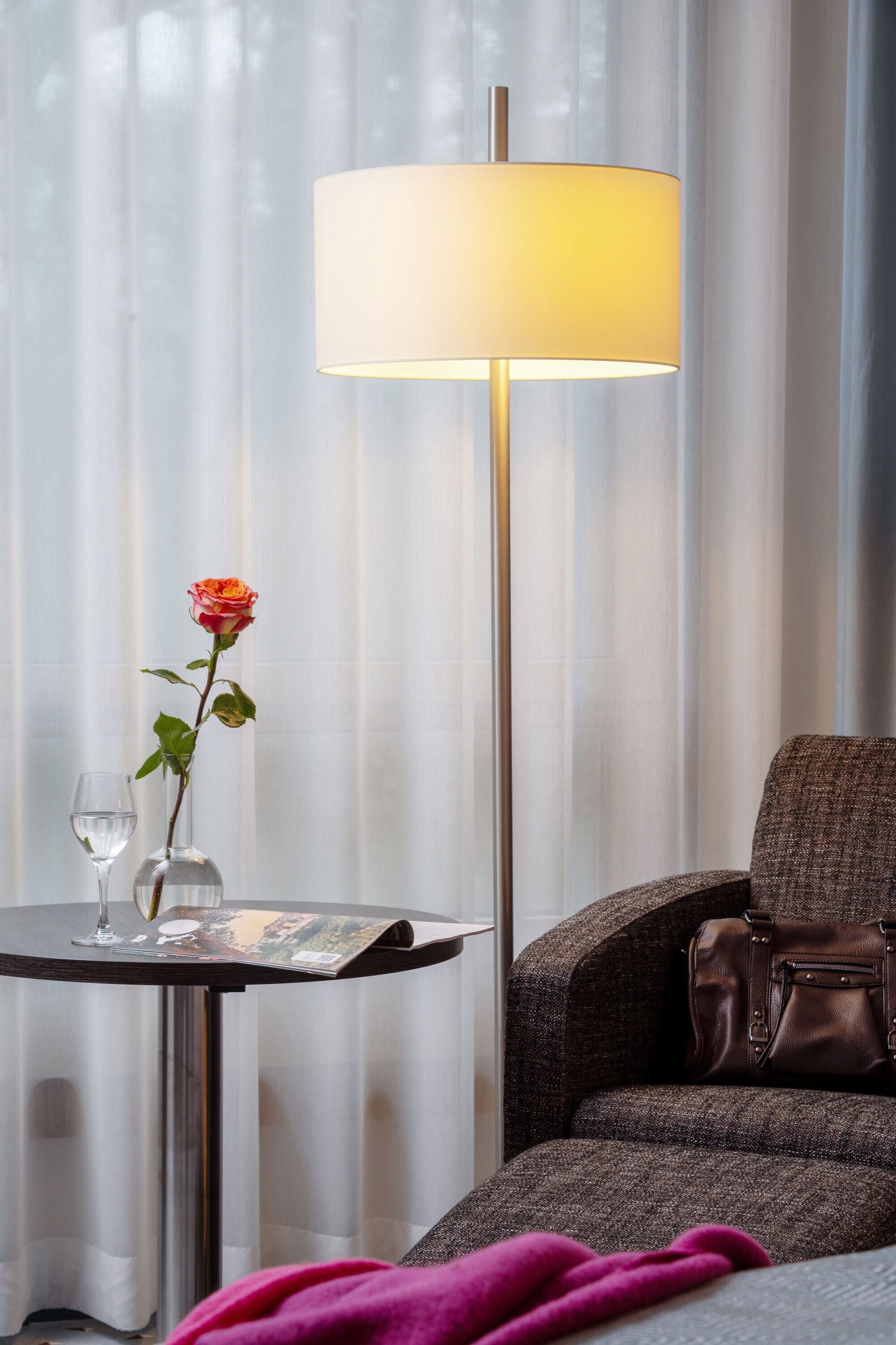 A hotel room with a table, a lamp, a chair, a rose, and a glass of water.