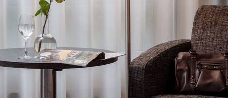 A hotel room with a table, a lamp, a chair, a rose, and a glass of water.