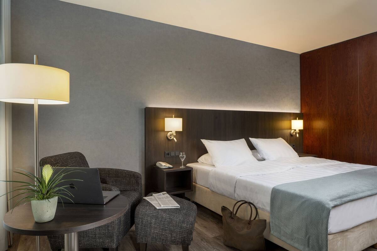 Ein Hotelzimmer mit Bett, Nachttisch, Lampe, Laptop, Stuhl und einem kleinen Tisch mit einer Pflanze.