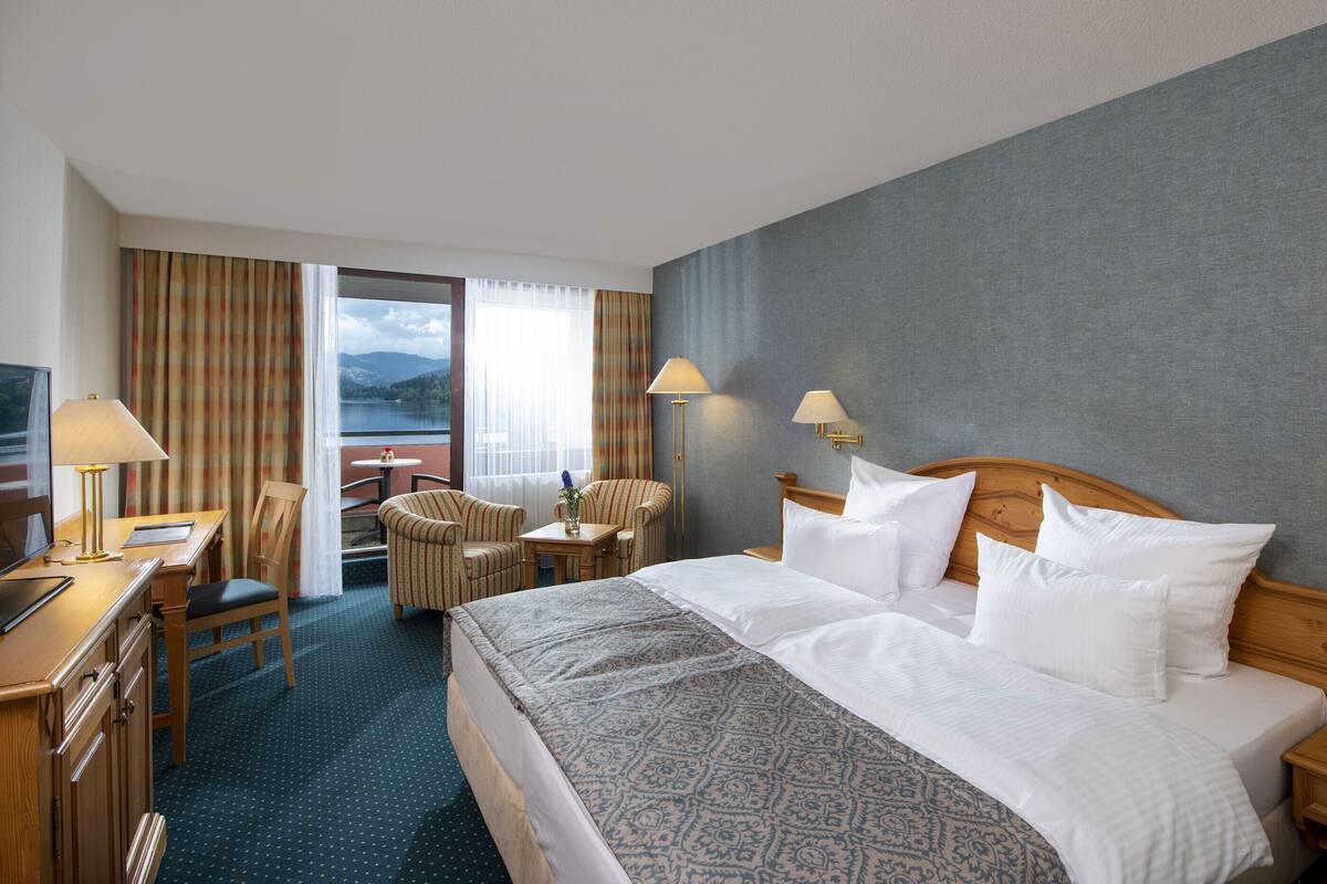 A hotel room with a large bed, two bedside lamps, a desk with a lamp, a chair, and a view of a lake through the window.