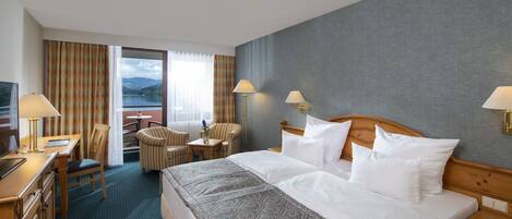 Ein Hotelzimmer mit einem großen Bett, zwei Nachttischlampen, einem Schreibtisch mit Lampe, einem Sessel und Blick auf einen See durch das Fenster.