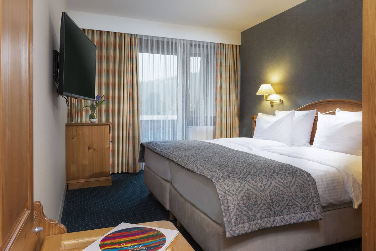 A hotel room with a bed, a TV, a desk, and a window with curtains.