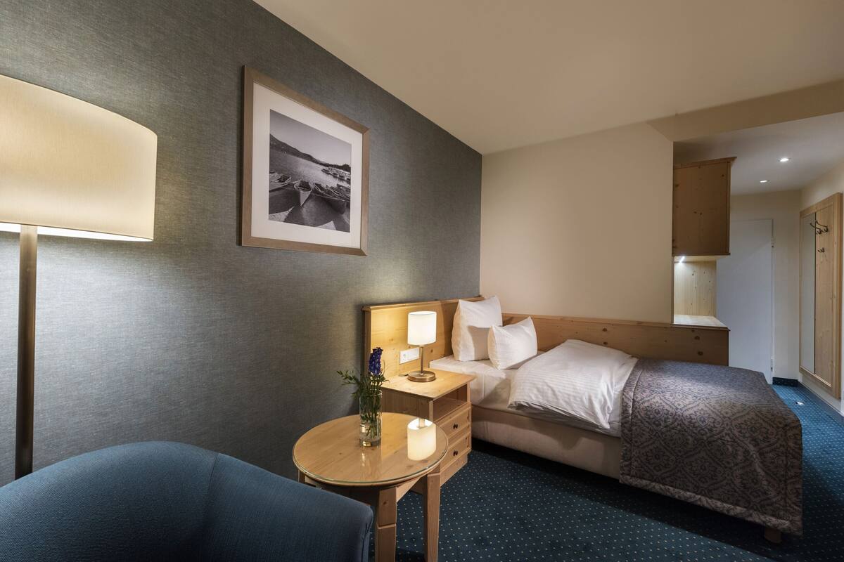 A hotel room with a bed, bedside table with a lamp, a chair, a small table with a candle, and a framed picture on the wall.
