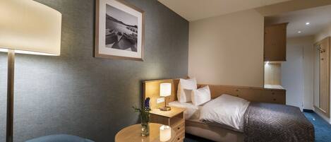 Une chambre d’hôtel avec un lit, une table de chevet avec une lampe, une chaise, une petite table avec une bougie et un tableau encadré au mur.