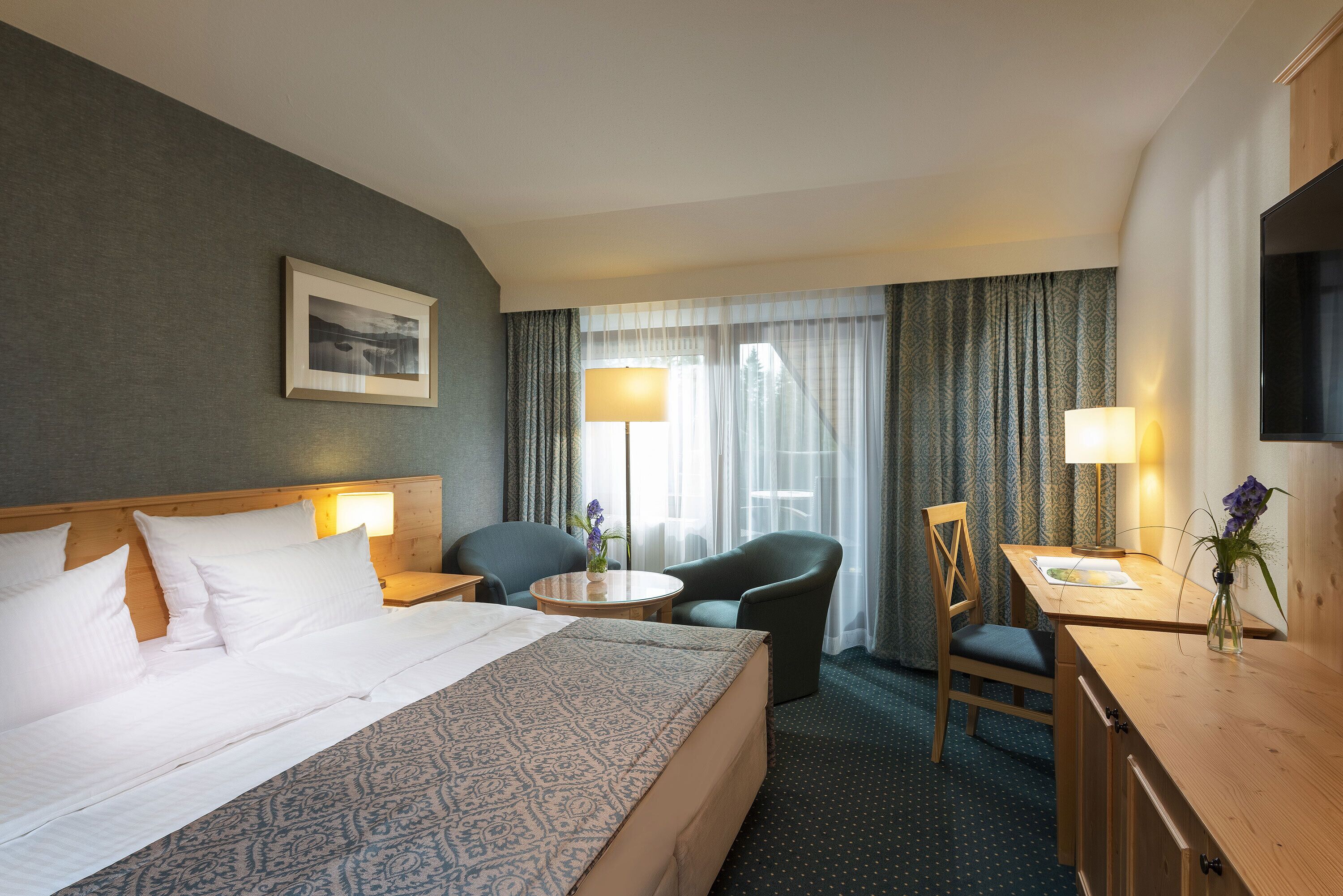 A hotel room with a large bed, two chairs, a desk, a TV, and a window with curtains.