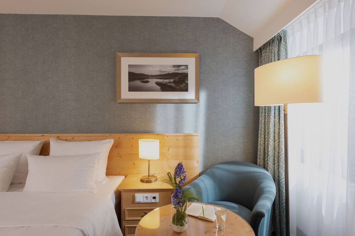 A hotel room with a bed, a nightstand with a lamp, a chair, a small table with a vase and book, and a framed picture on the wall.