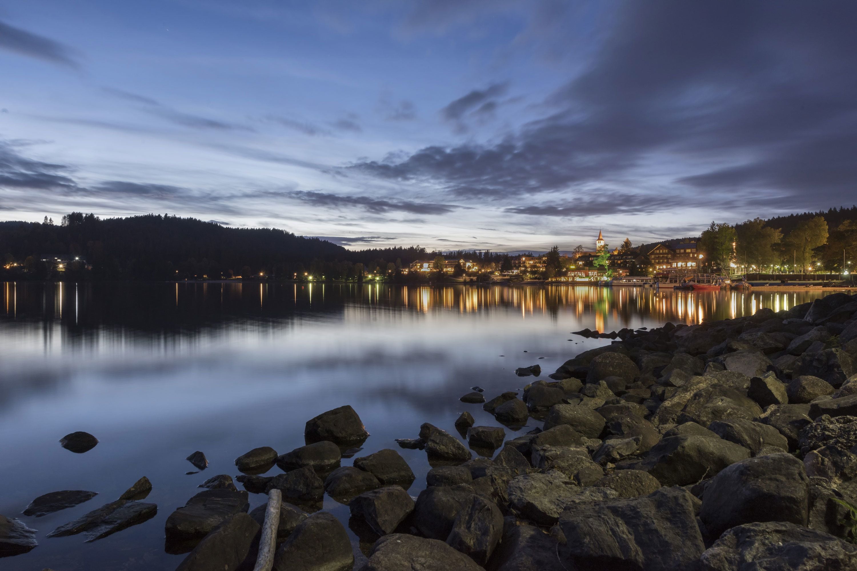 A lakeside view at dusk with calm waters reflecting lights from buildings and trees.
