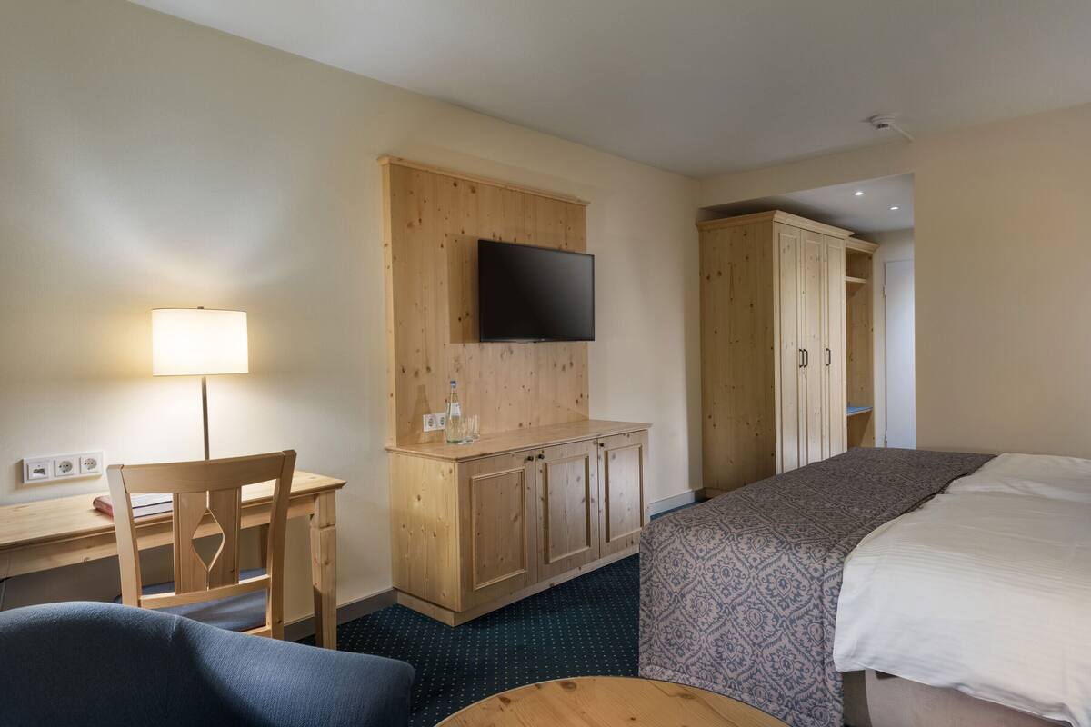 A hotel room with a bed, a desk with a chair, a TV mounted on the wall, and a wardrobe.