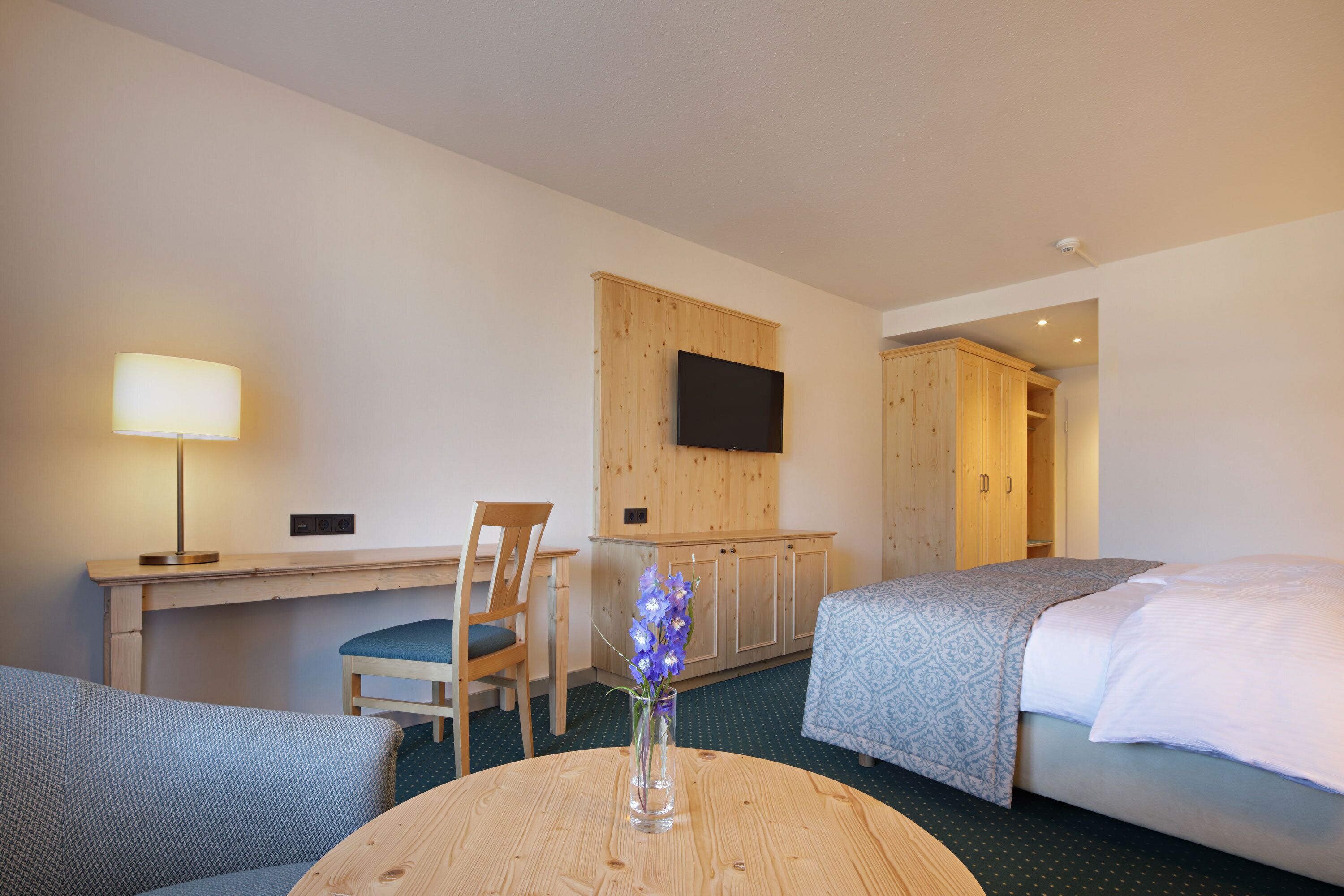 Una habitación de hotel con una cama, un escritorio con lámpara, un televisor montado en la pared y un armario de madera.