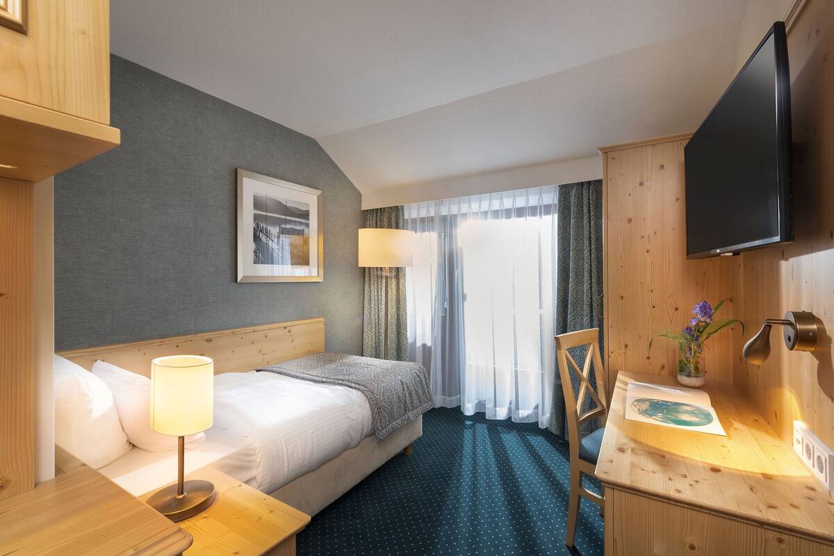 A hotel room with a bed, a desk with a lamp, a television, and a window with curtains.