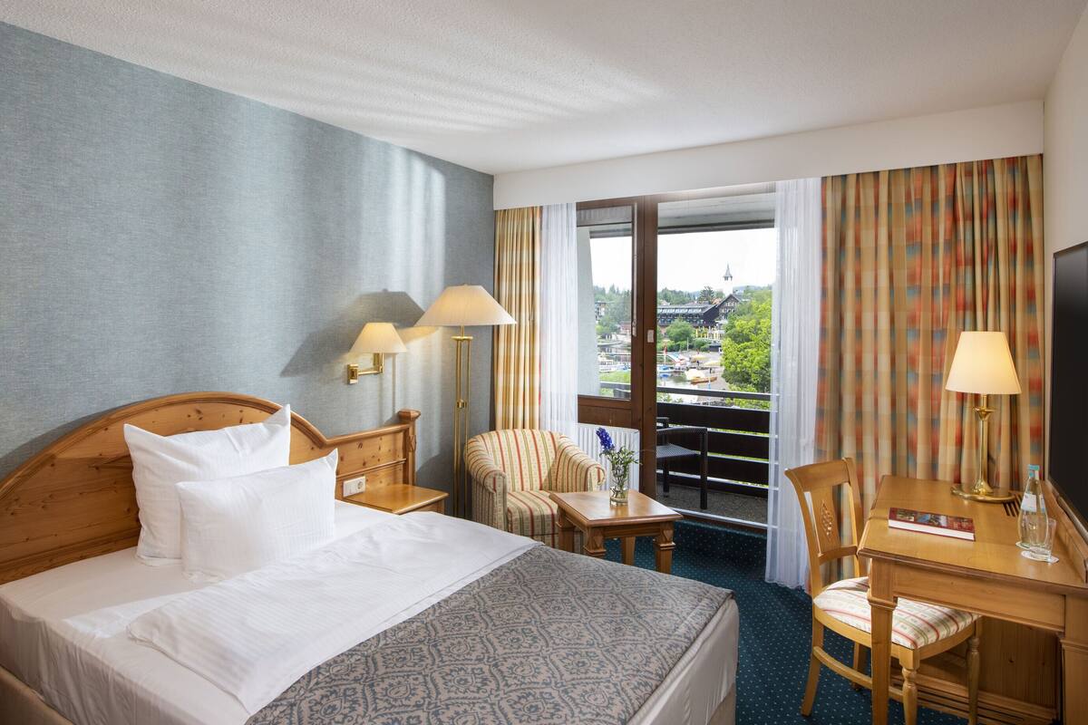A hotel room with a large bed, a wooden desk, two chairs, a small table, and a view of a cityscape through the window.