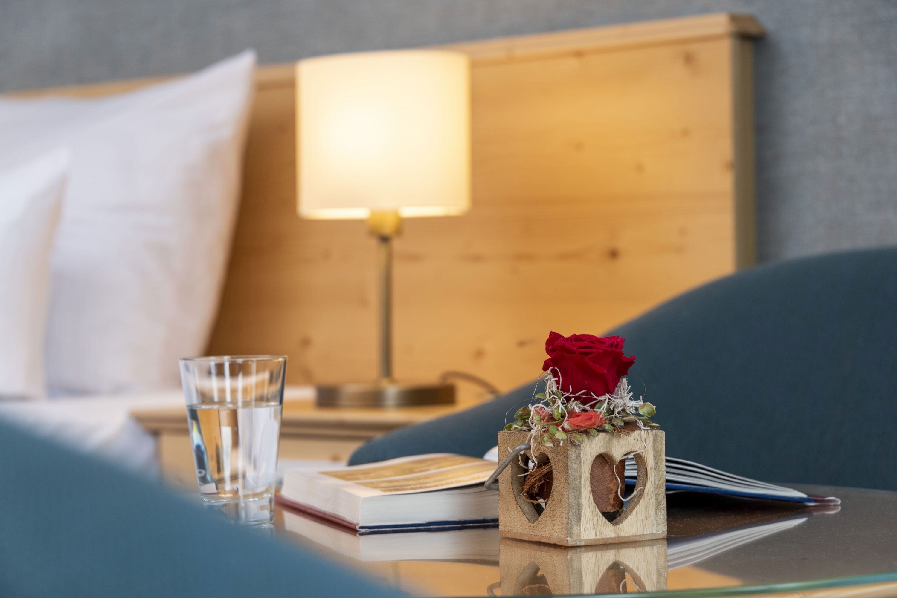 Habitación de hotel impecable con una cama, una mesita de noche con un vaso de agua, un libro y un bloque de madera decorativo con una rosa roja.