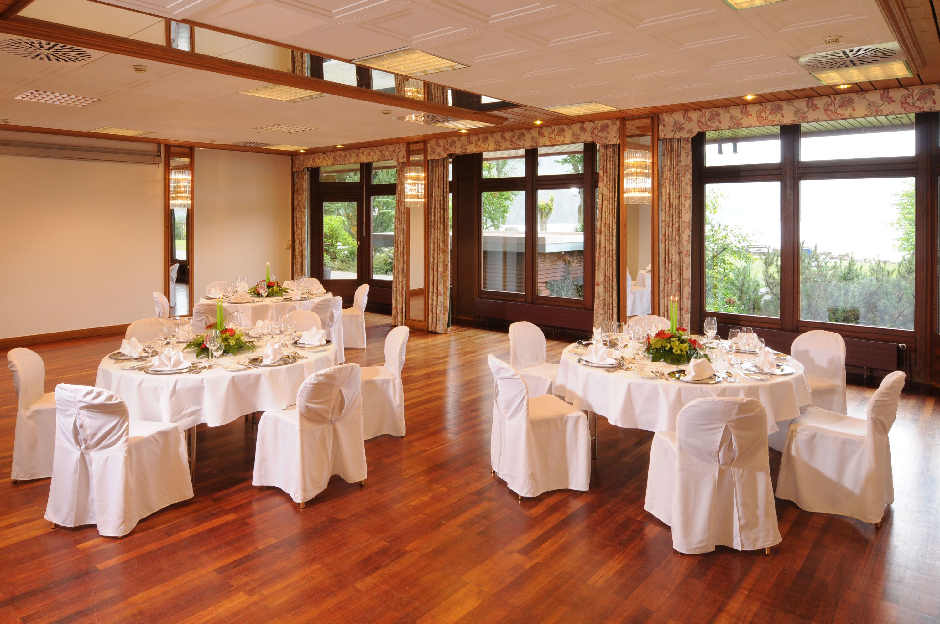 A large conference room with round tables set for a formal event, large windows with curtains, and wooden flooring.