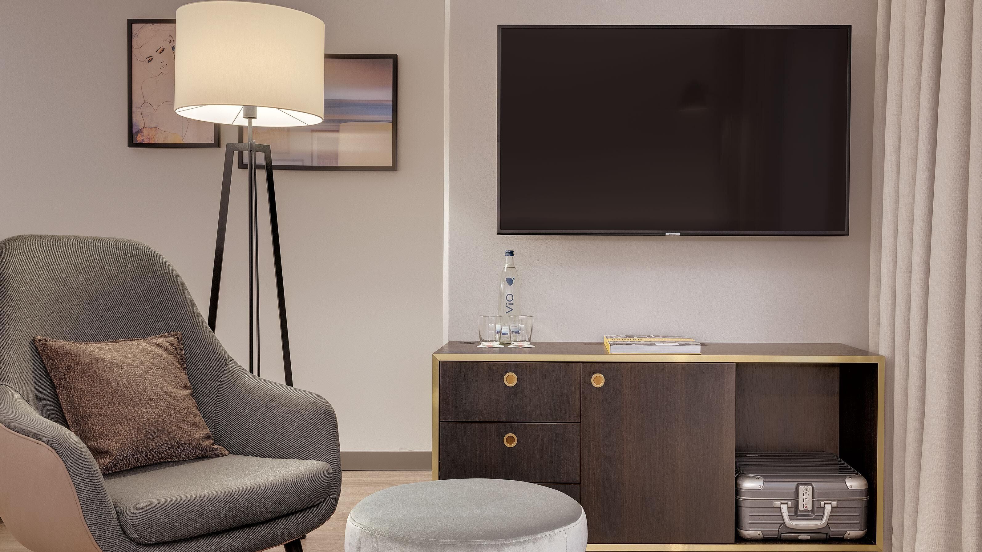 A modern hotel room with a grey armchair, a floor lamp, a TV mounted on the wall, a small wooden side table with a book, and a suitcase on the floor.