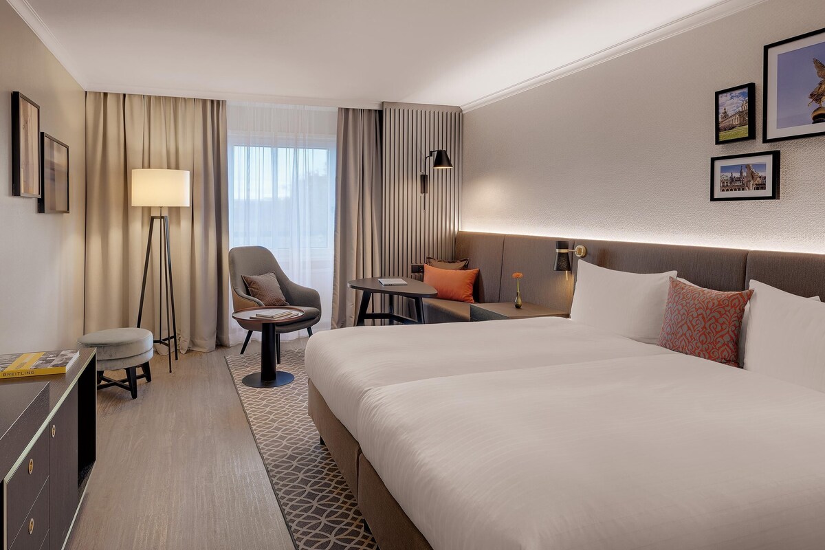 A hotel room with a large bed, a desk with a chair, a sofa, a small table with a lamp, and a TV.