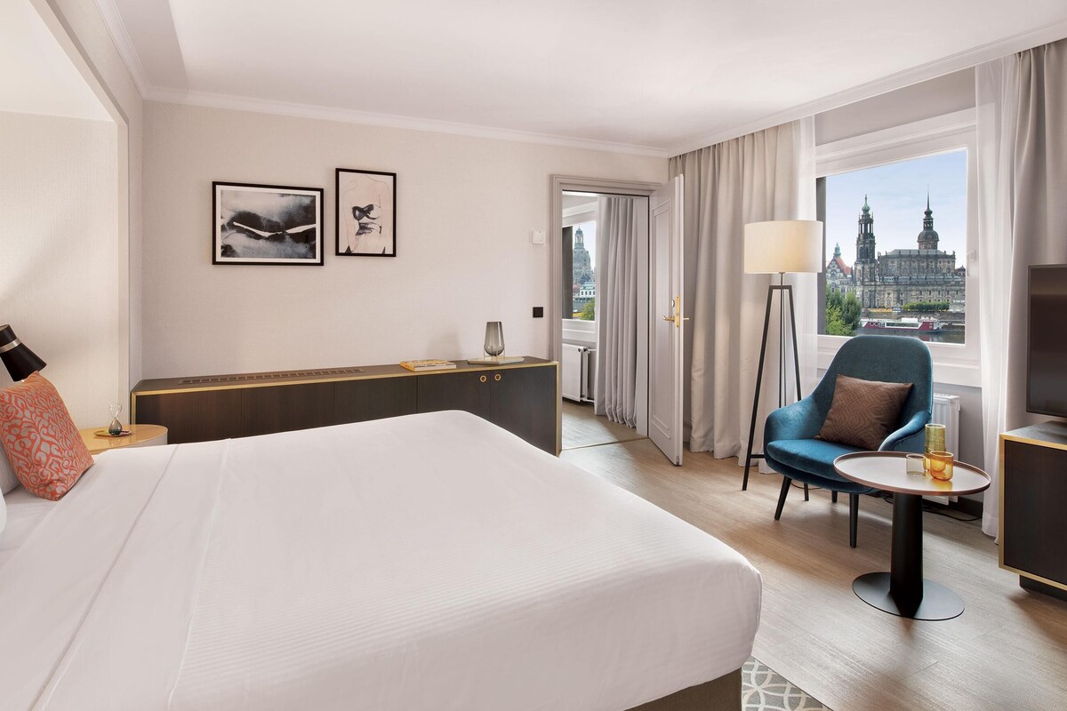 A hotel room with a large bed, a desk, a chair, a TV, and a window with a city view.