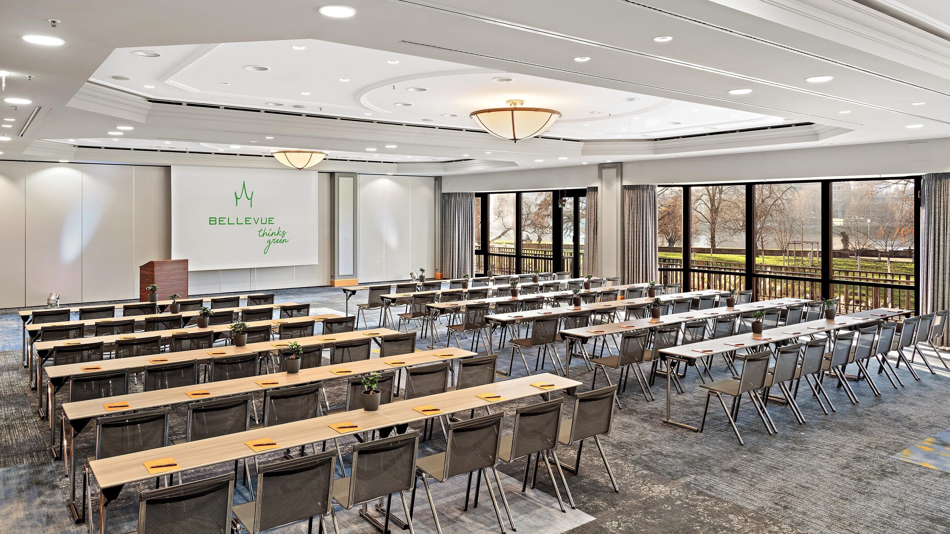 A conference room with rows of tables and chairs, a projector screen, and a large window with a view of a park.