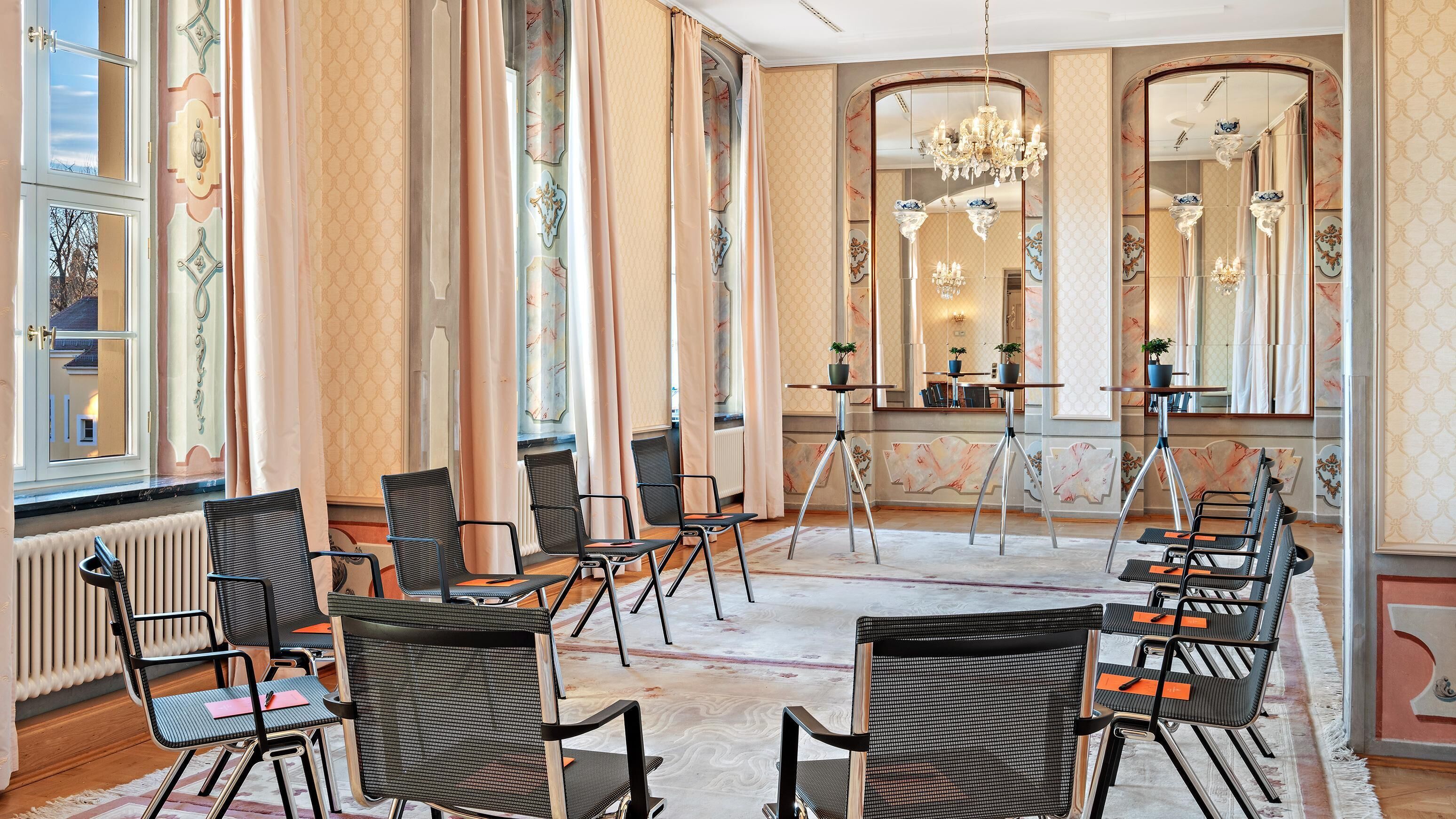 A spacious room with large windows, a chandelier, and multiple chairs arranged for seating.
