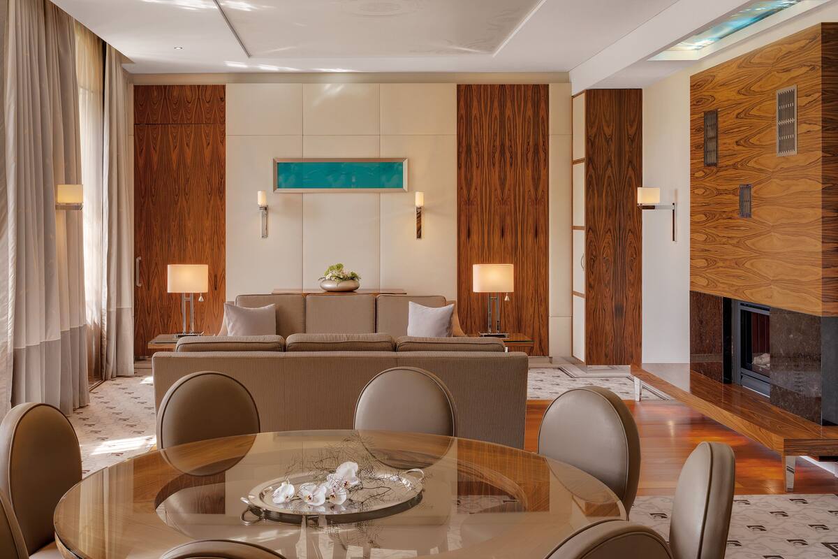 A modern hotel room with a central seating area, a glass dining table with chairs, and a fireplace.