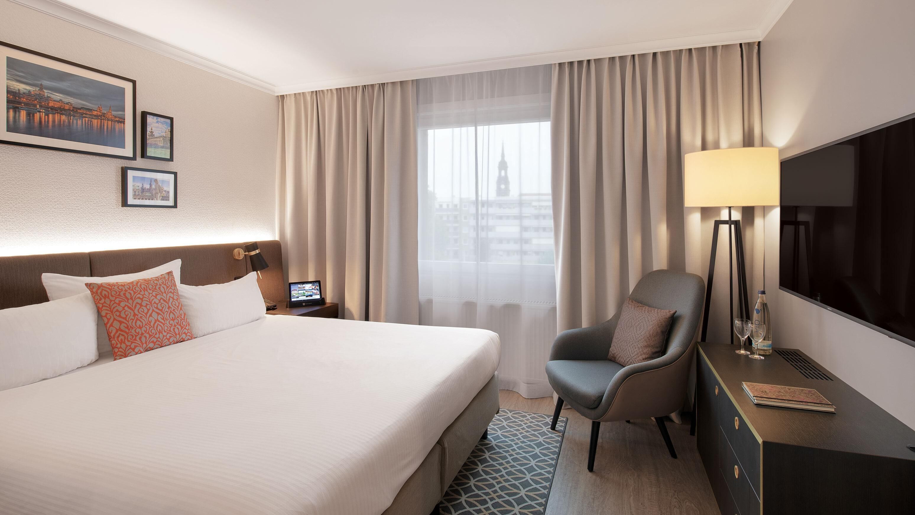 A hotel room with a large bed, a desk with a lamp, a chair, a TV, and a window with curtains.