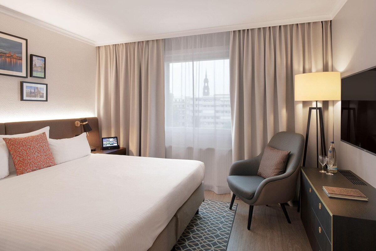 A hotel room with a large bed, a desk with a lamp, a chair, a TV, and a window with curtains.