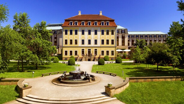 A large, historic building with a central courtyard, a fountain, and surrounding greenery.