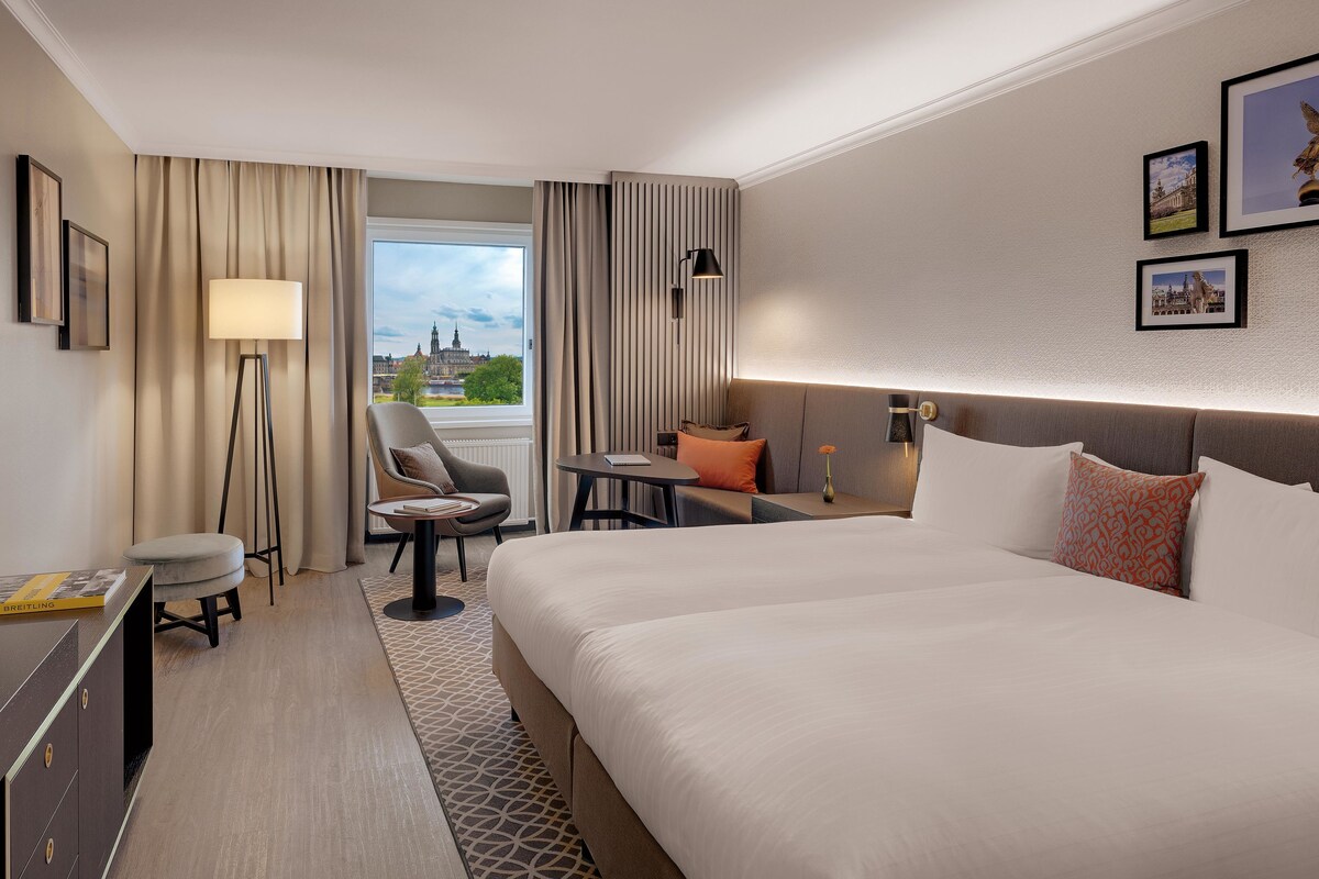 A hotel room with a large bed, a desk, a chair, a small table, and a TV.