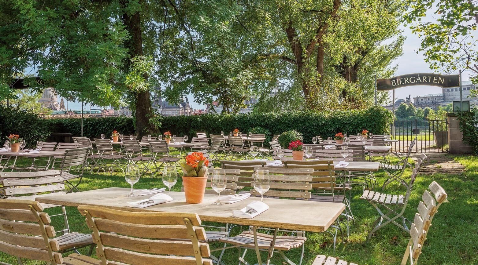 Un espacio de comedor al aire libre con mesas y sillas, rodeado de vegetación y vistas a un paisaje urbano.