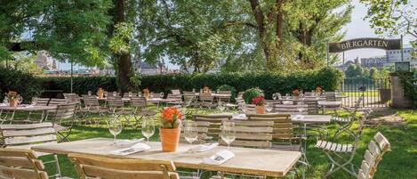 Un espacio de comedor al aire libre con mesas y sillas, rodeado de vegetación y vistas a un paisaje urbano.