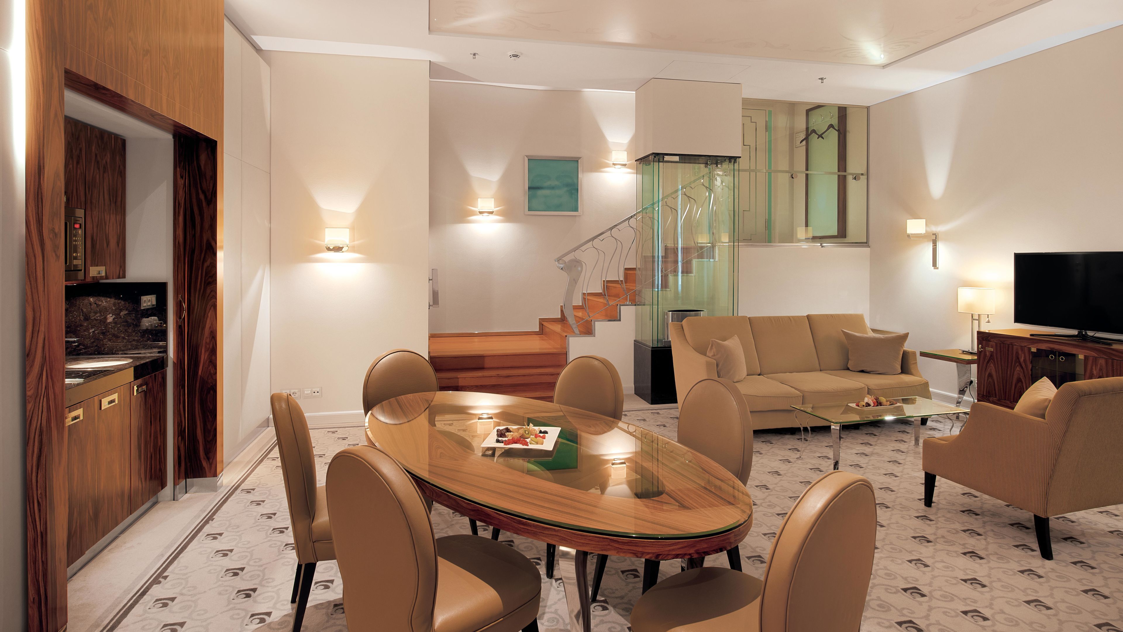 A modern living room with a glass dining table, beige sofas, a staircase, and a TV.