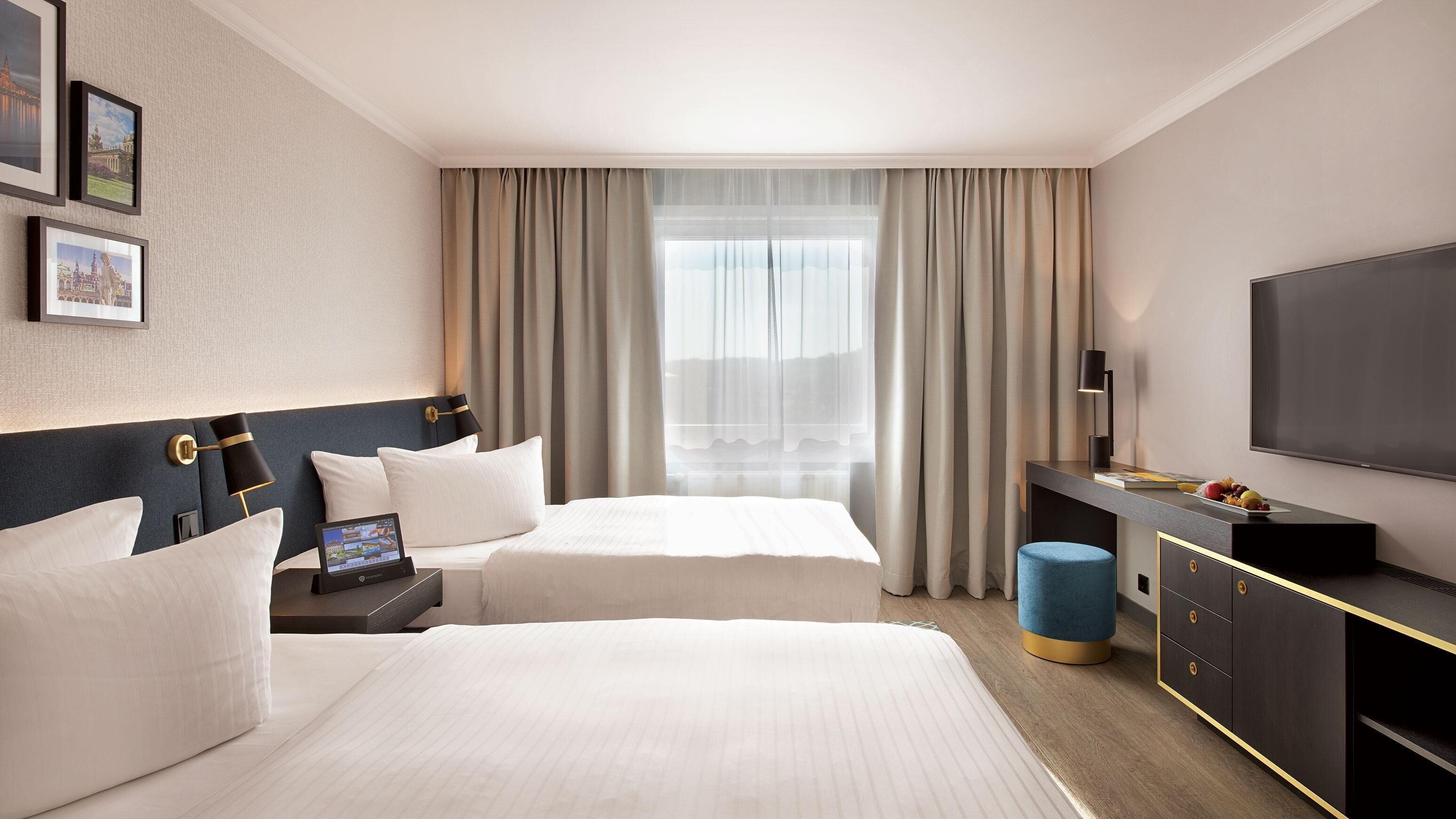 Une chambre d’hôtel avec deux lits, un bureau avec une télévision, une lampe et un petit tabouret bleu.