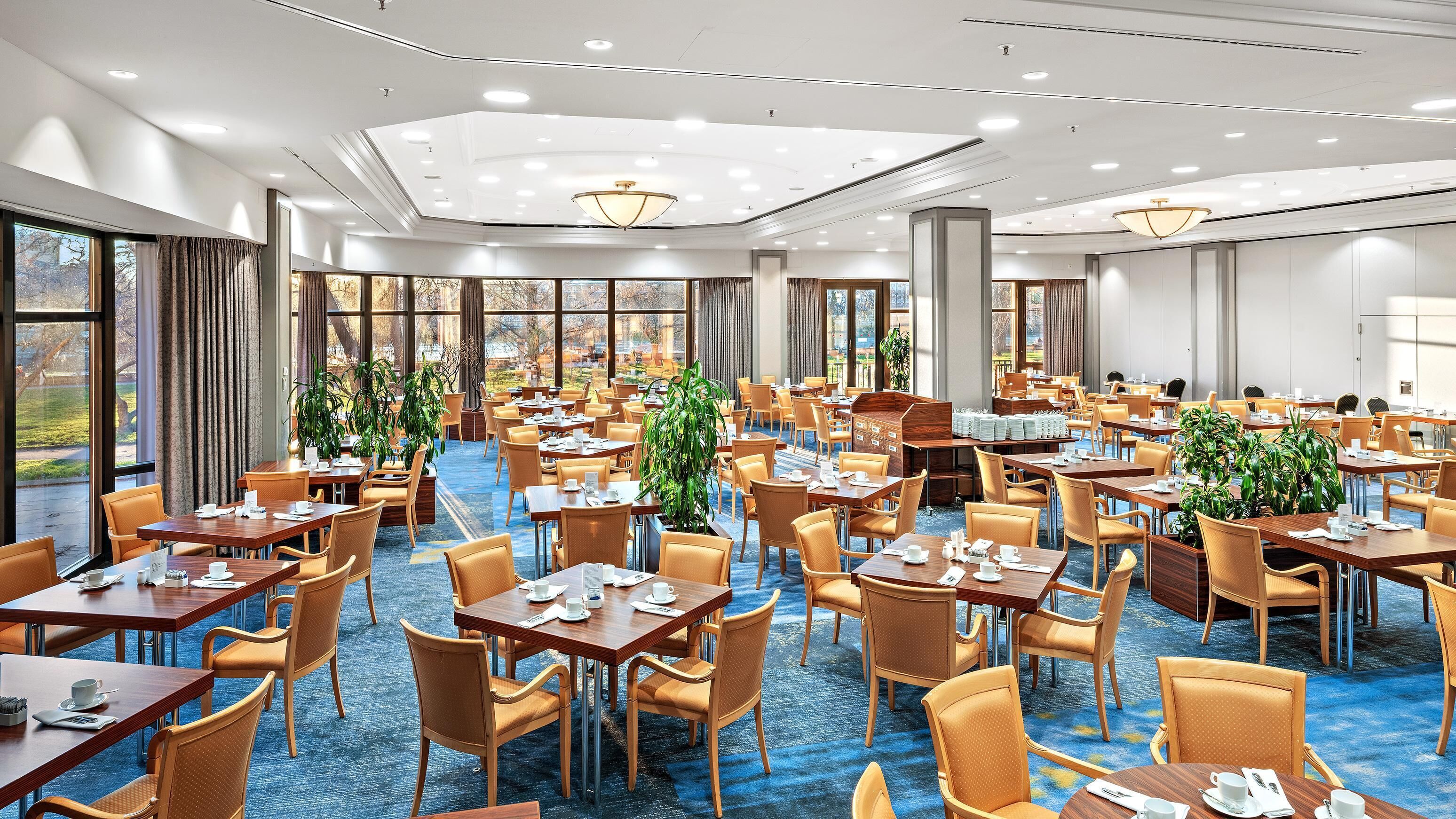 A hotel dining area with multiple tables and chairs, large windows, and ceiling lights.