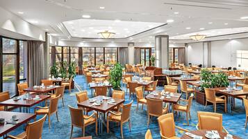 A hotel dining area with multiple tables and chairs, large windows, and ceiling lights.