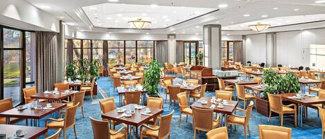 Comedor de hotel con varias mesas y sillas, ventanas amplias e iluminación en el techo.