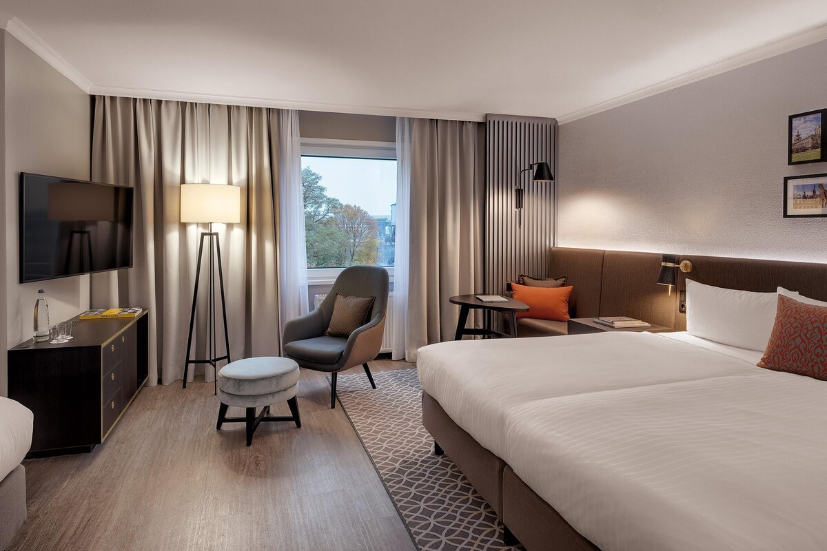 A hotel room with a large bed, a desk with a TV, a chair, a floor lamp, and a view of the outdoors.