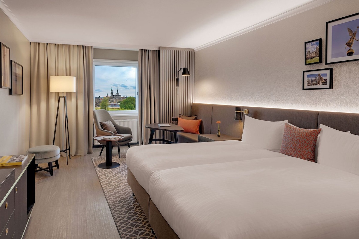 Habitación de hotel con cama, escritorio, silla, lámpara y vista a un paisaje urbano.