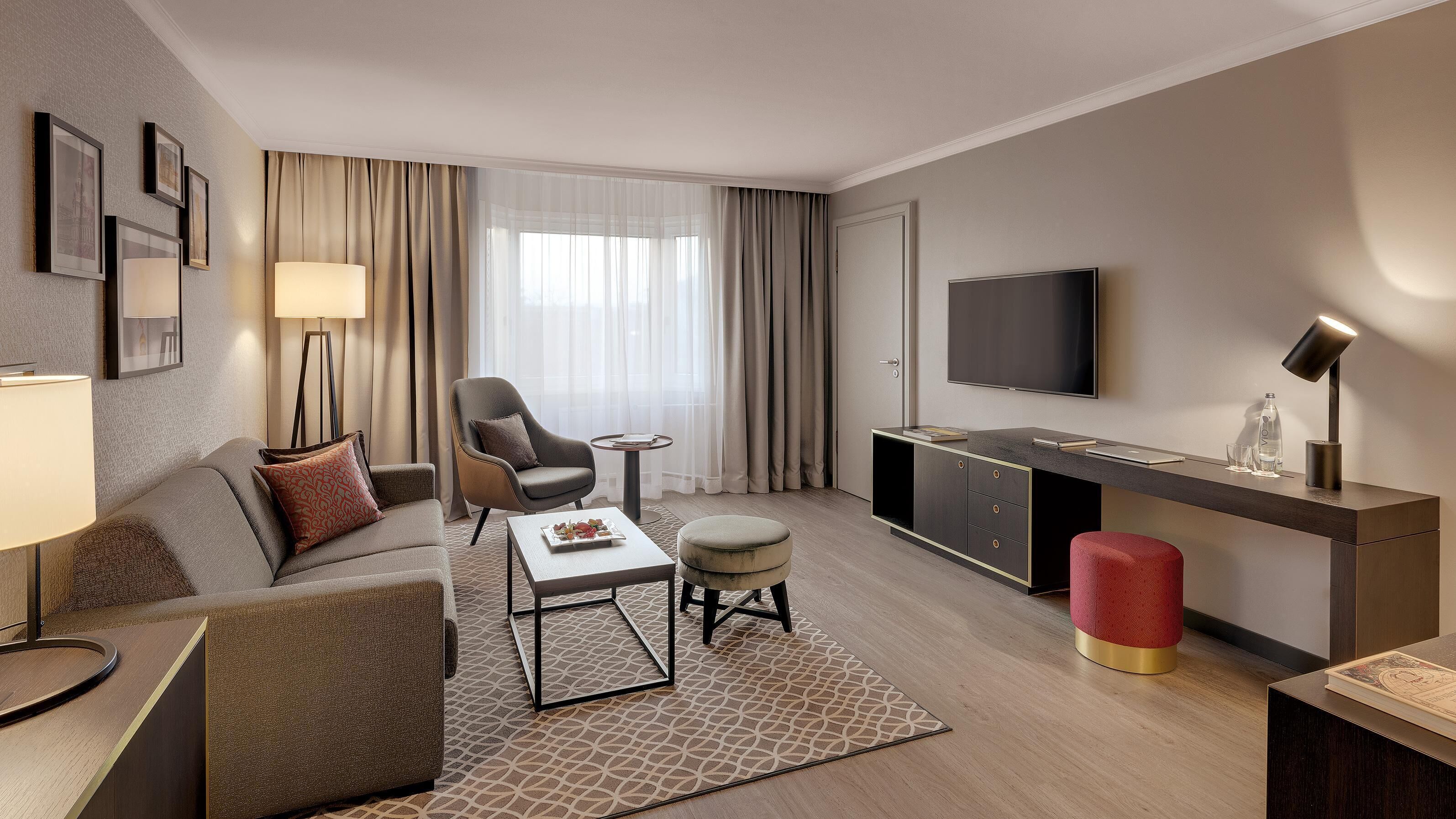 Une chambre d’hôtel moderne équipée d’un canapé, d’un fauteuil, d’une table basse, d’un pouf et d’un bureau avec un téléviseur à écran plat.