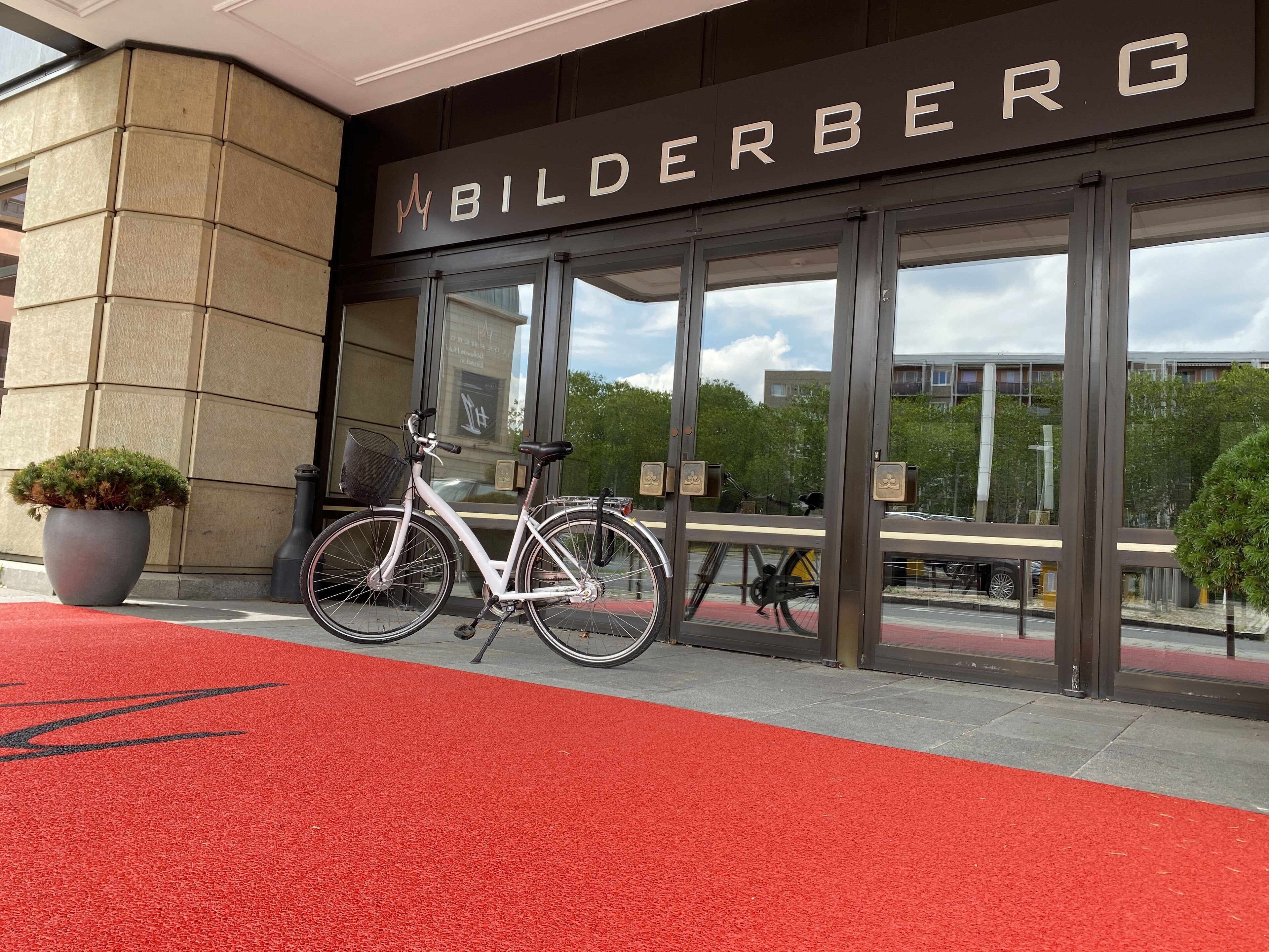 A hotel entrance with a red carpet, a white bicycle, and a large planter.