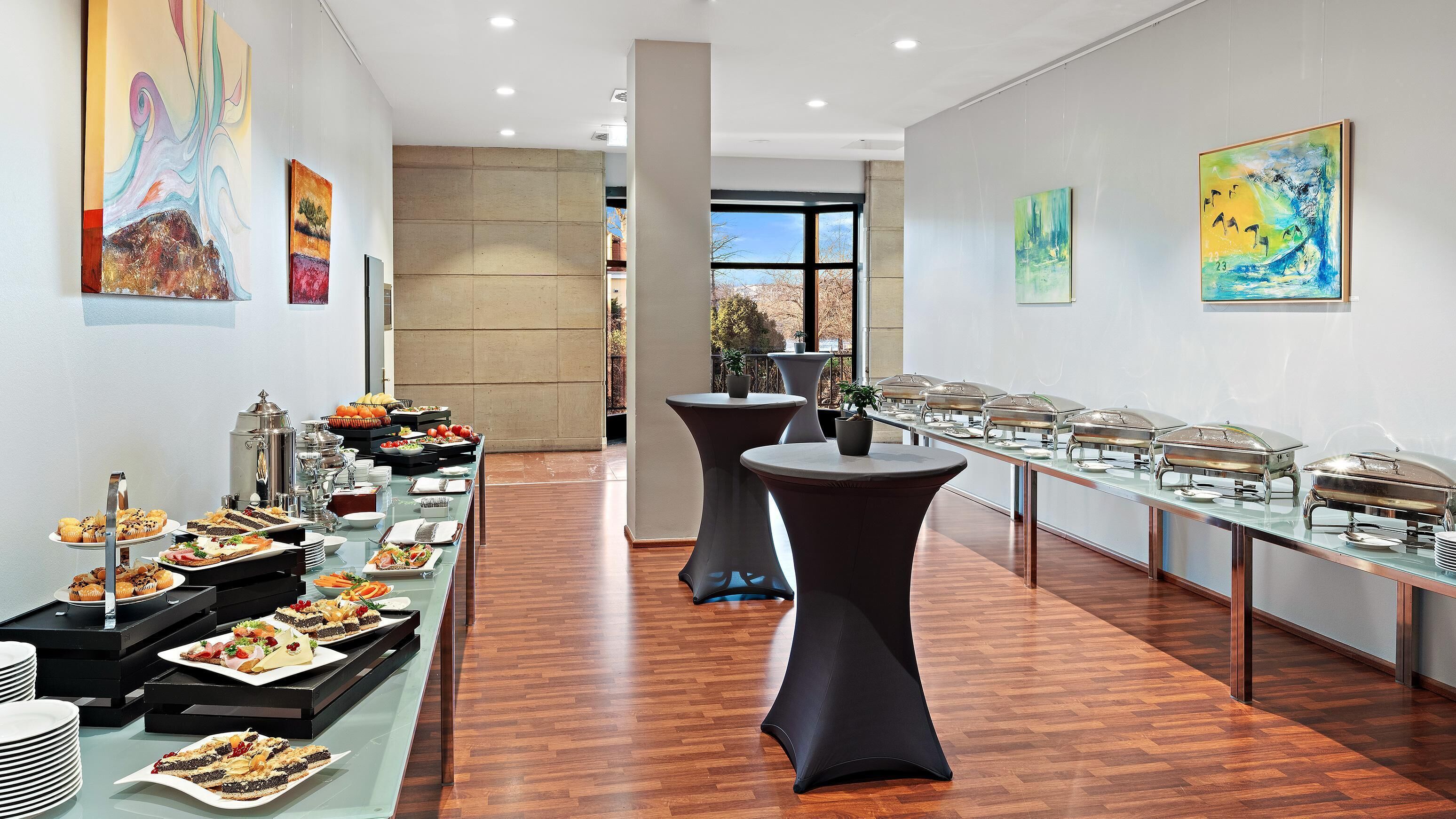 A buffet setup with various food items and buffet covers, with artwork on the walls and a view of the outdoors.