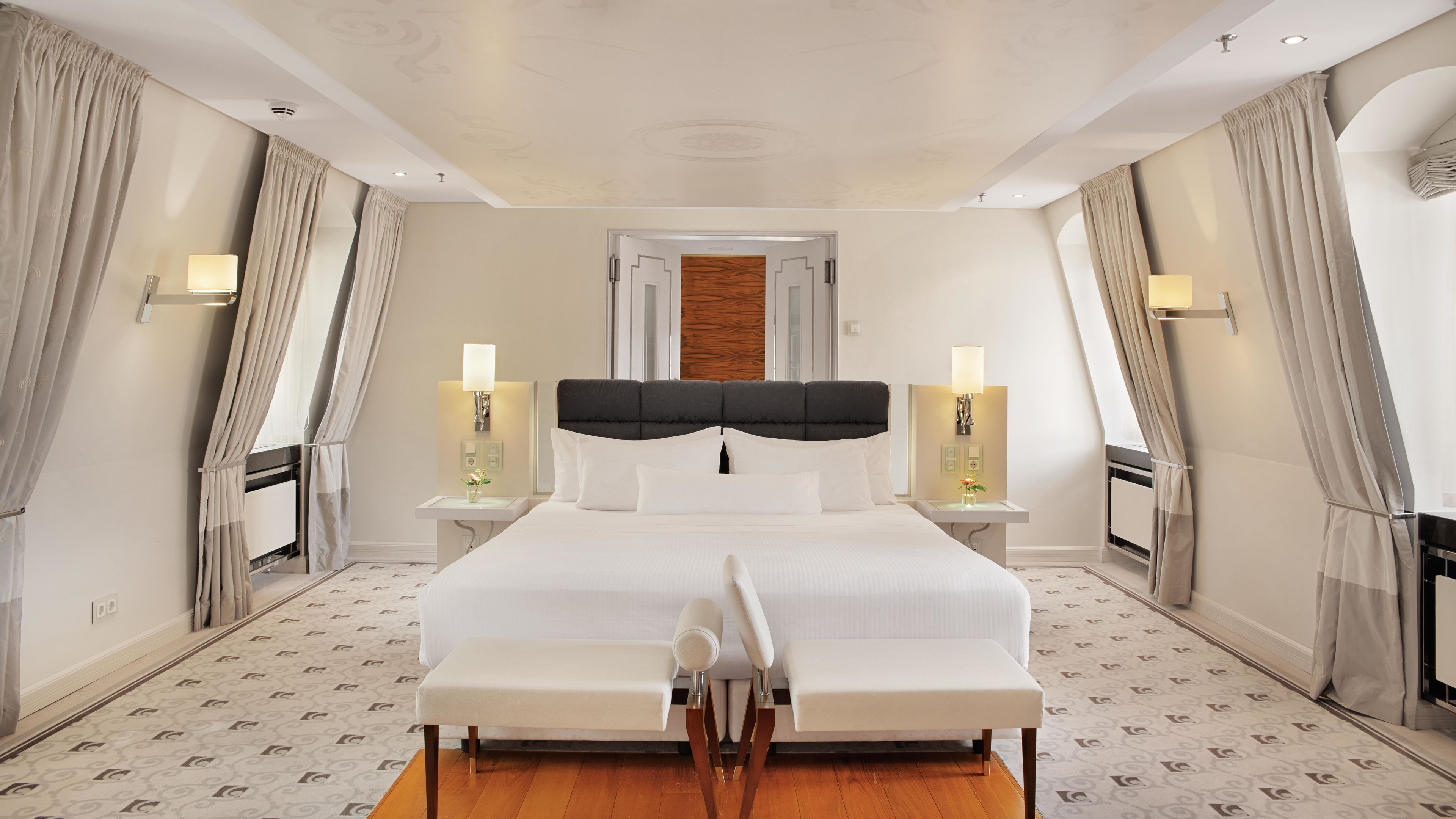 A modern hotel room with a large bed, two bedside tables with lamps, a wooden headboard, and a patterned floor.