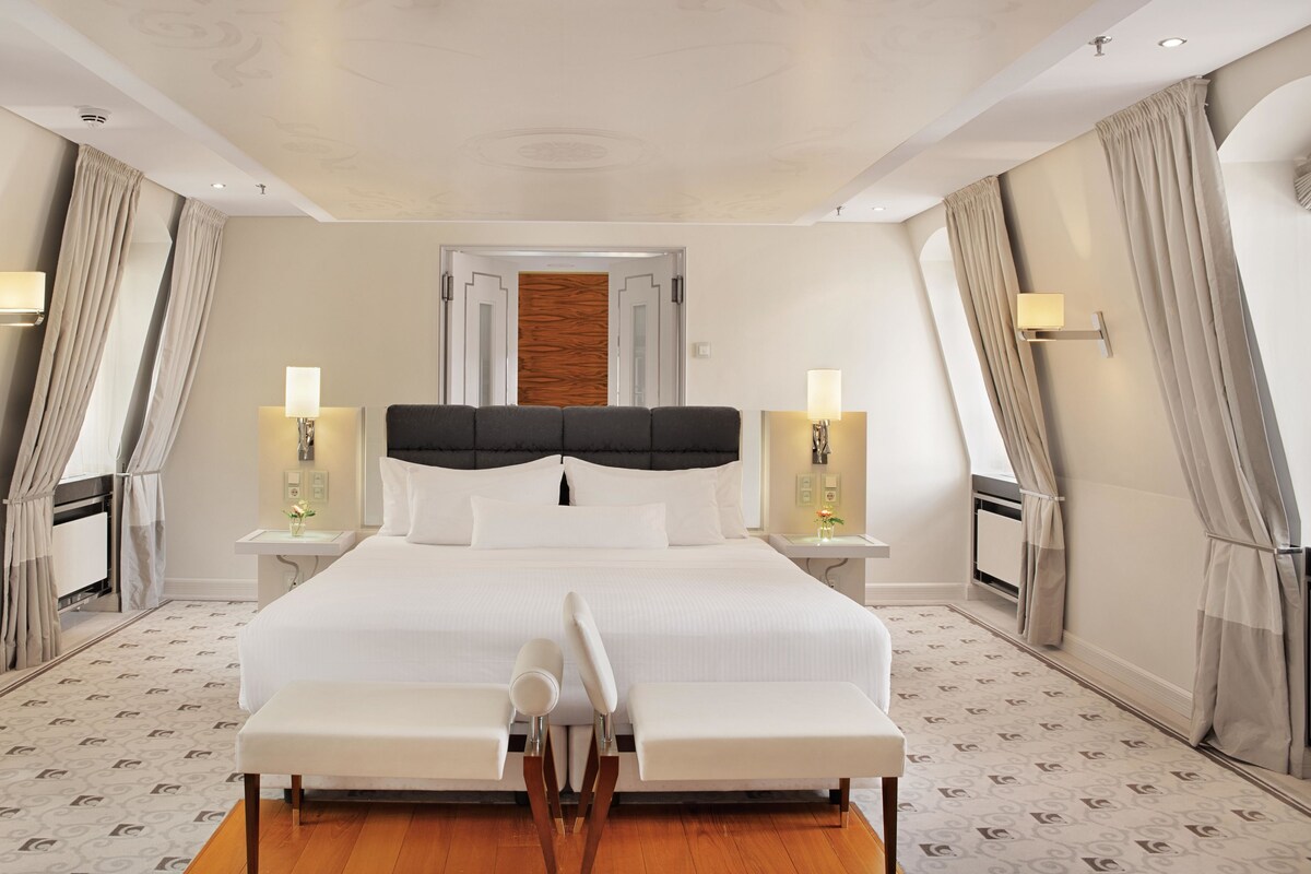 A modern hotel room with a large bed, two bedside tables with lamps, a wooden headboard, and a patterned floor.