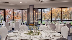A banquet hall with round tables set for a formal event, large windows offering a view of autumn trees, and a candelabra centerpiece.